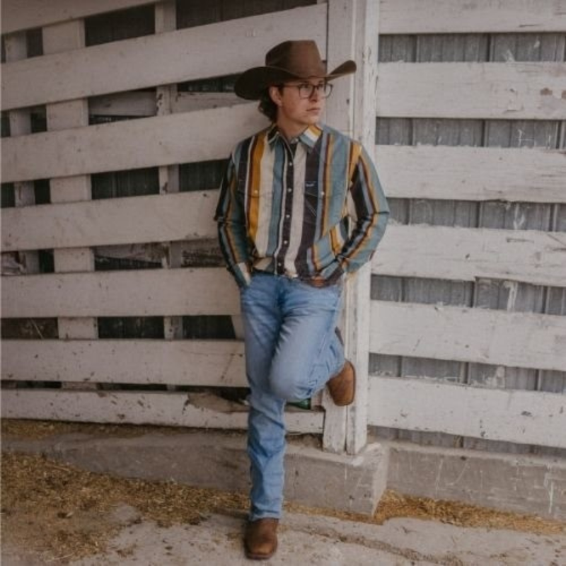 Man wearing a cowboy hat and striped shirt leaning against a wooden fence.