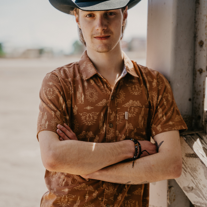 Man wearing a patterned shirt and cowboy hat standing with arms crossed.